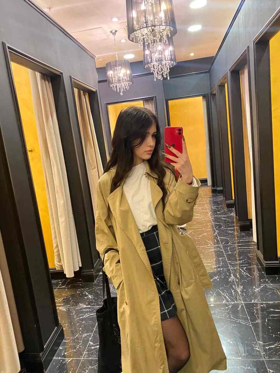 Mujer toma una selfie en el espejo de un pasillo con mármol, usando un teléfono rojo, un abrigo color caqui y una falda a cuadros.