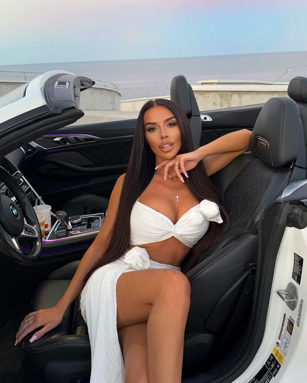 A woman in a white two-piece outfit sits in a white convertible car with the ocean behind her.