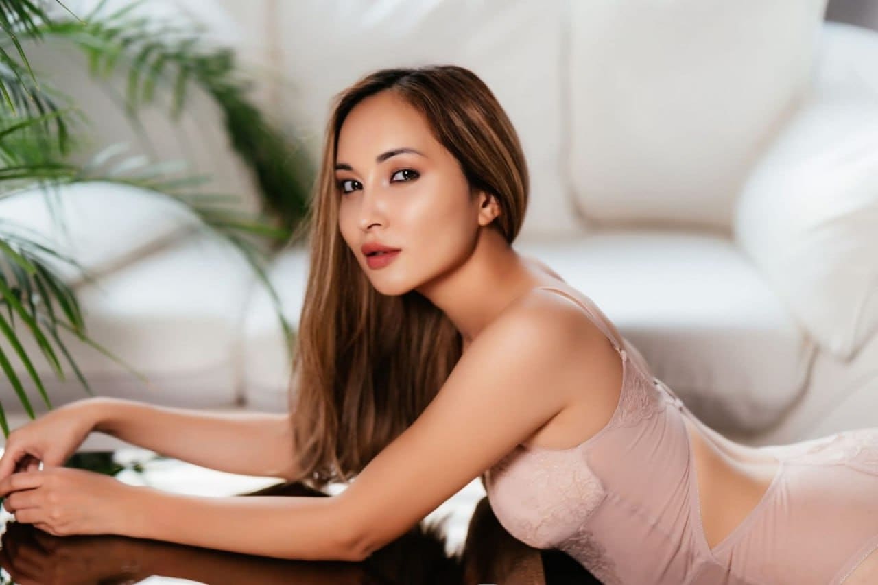 A woman in a pink lace bodysuit leans forward on a dark table, set against a white sofa and green plant leaves.