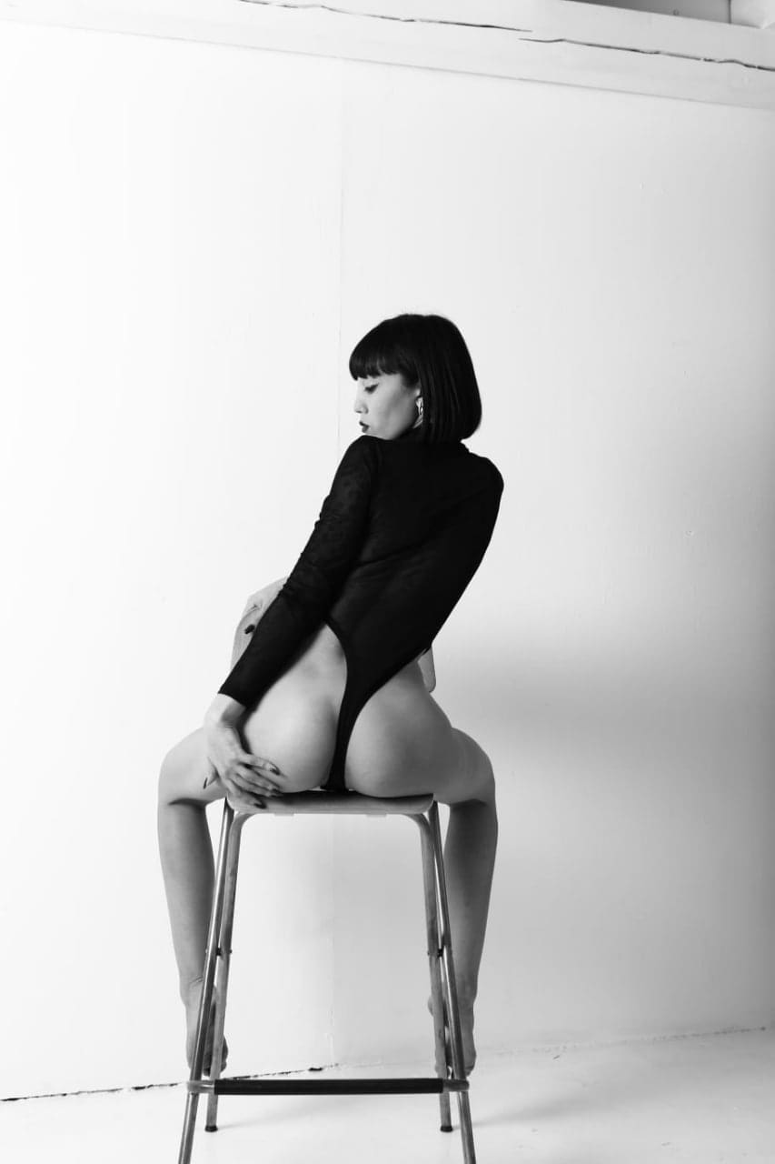 Mujer con bob oscuro en taburete metálico sobre fondo blanco, lleva bodysuit negro de manga larga con abertura frontal alta que expone el torso.