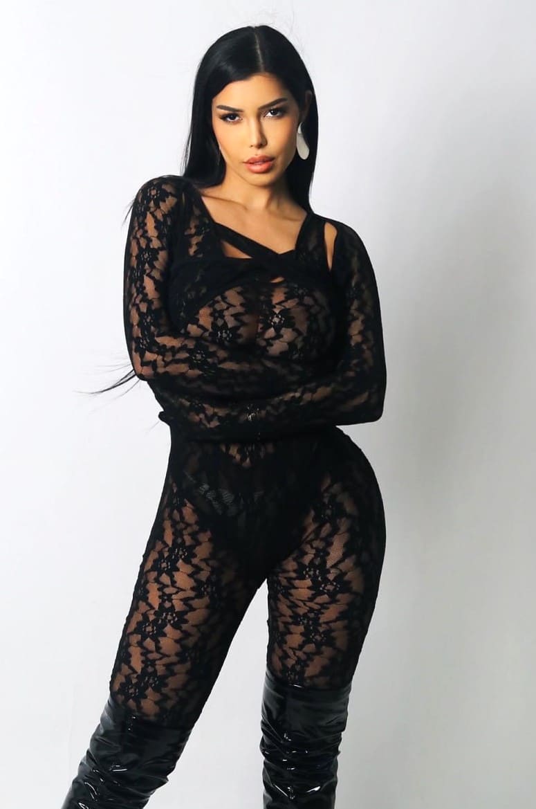 Mujer de pie con brazos cruzados, body negro de encaje transparente y botas altas negras sobre fondo blanco.