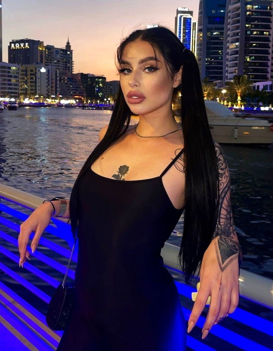 Aleks en un paseo marítimo al atardecer, con vestido negro de tirantes y choker, frente a una ciudad iluminada.