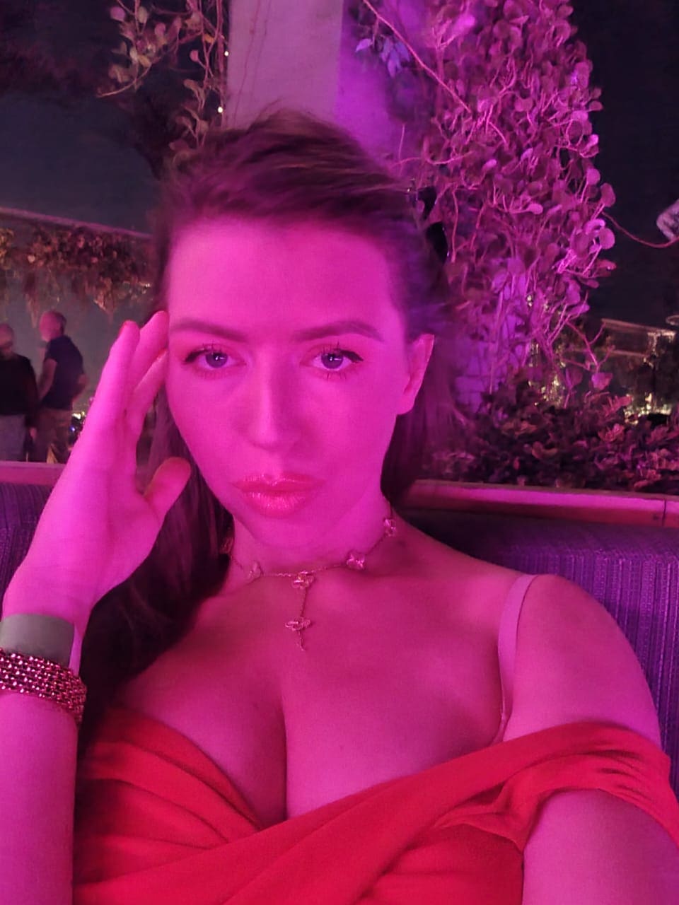 Mujer de cabello castaño recogido sentada en exterior nocturno, con vestido rojo de tirantes finos bajo luz rosa y morada.