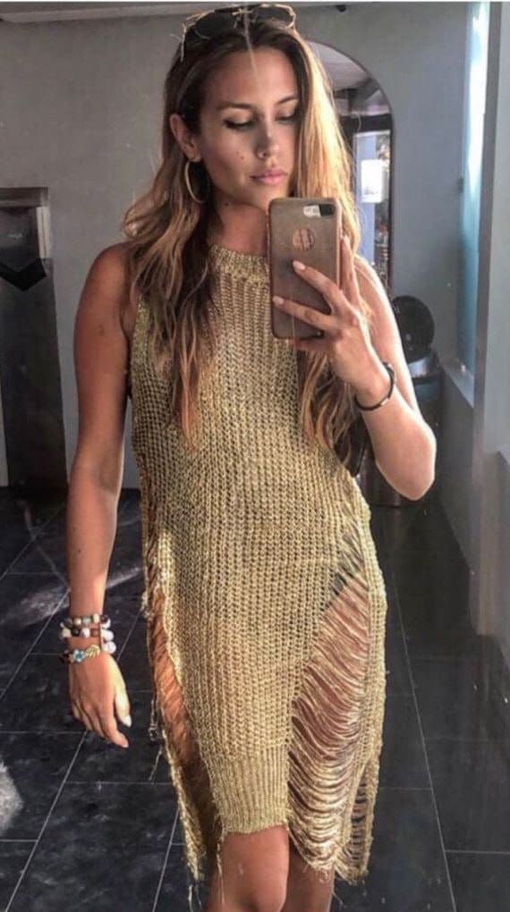 Mujer con gafas en la cabeza toma una selfie al espejo con vestido beige de punto abierto y cortes laterales, sobre suelo oscuro.