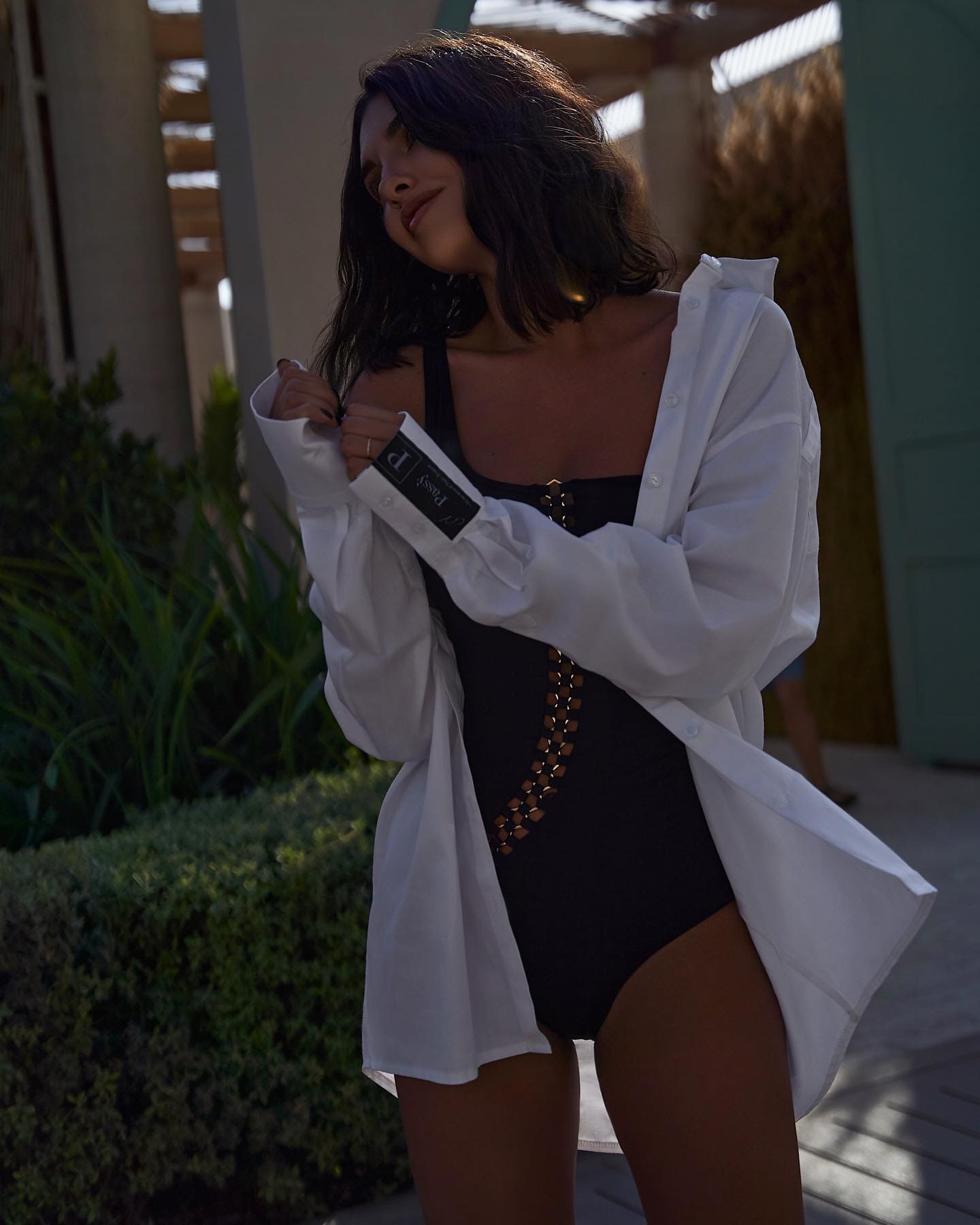 Mujer con cabello oscuro hasta los hombros al aire libre frente a columnas y vegetación, llevando un bañador negro con detalle dorado y una camisa blanca abierta sobre él.
