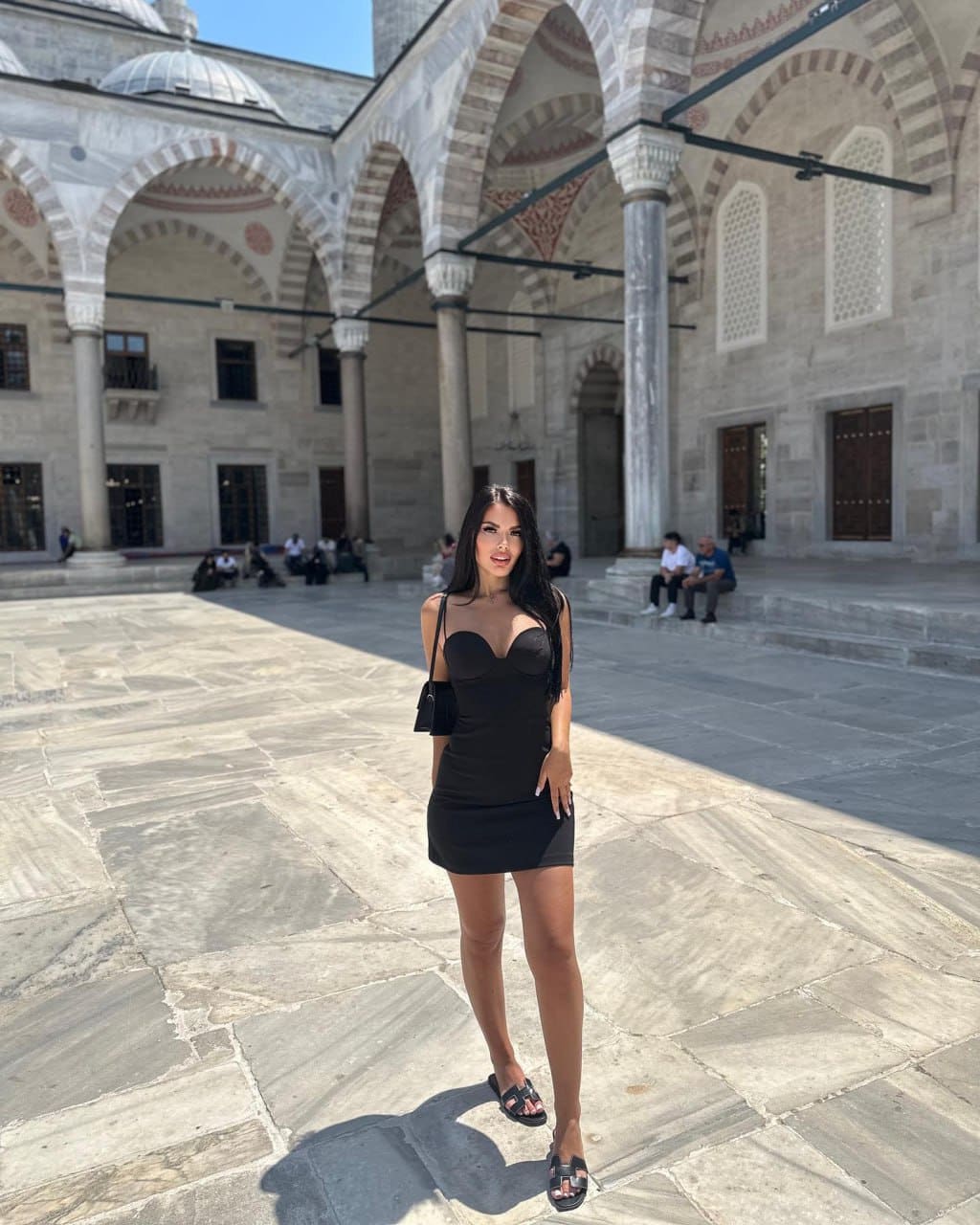 Mujer de pelo largo oscuro de pie en patio de piedra frente a edificio con arco, vestido negro sin mangas y bolso pequeño al hombro.