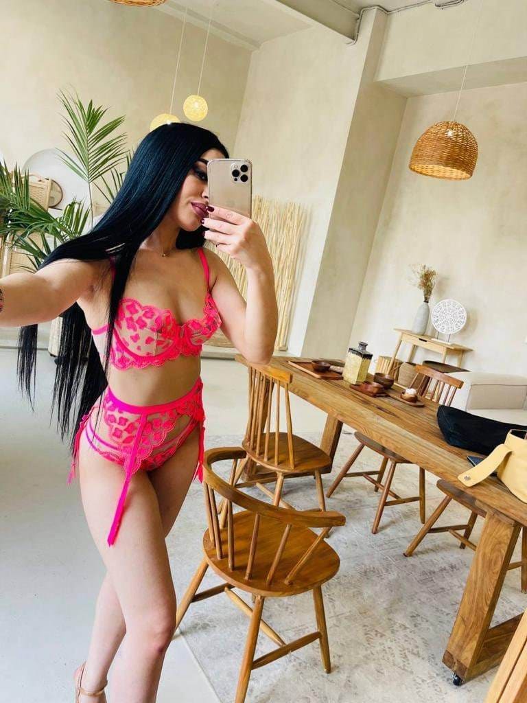 Donna con capelli scuri che scatta un selfie allo specchio, indossa un set di lingerie rosa in pizzo in una stanza con sedie di legno.