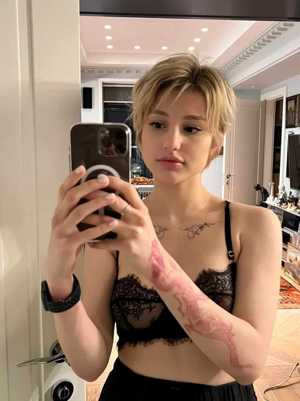 Mujer con cabello rubio corto usando lencería negra de encaje, mostrando tatuajes en el antebrazo izquierdo en una habitación interior bien iluminada.