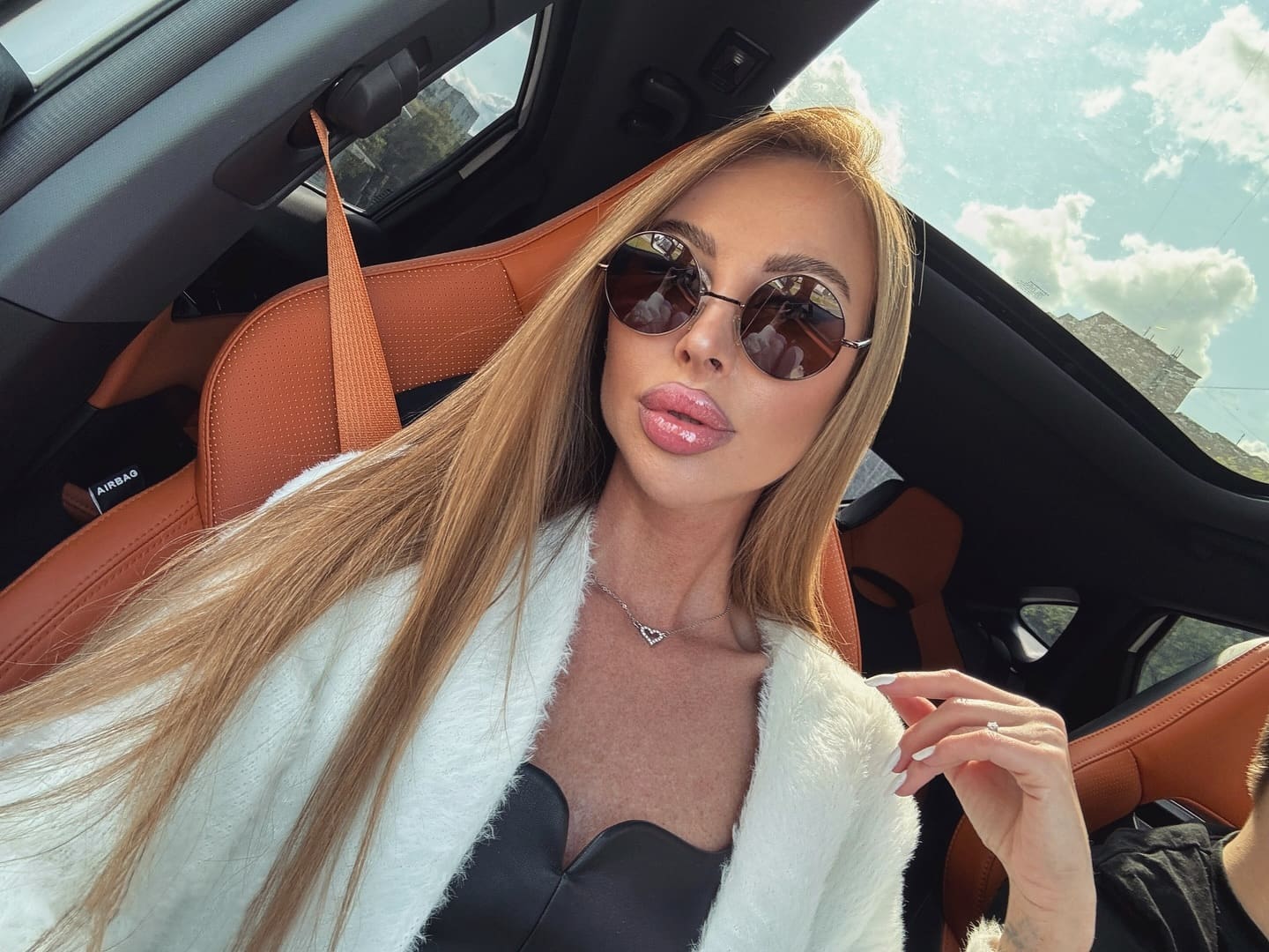 Mujer rubia con gafas redondas, cárdigan blanco peludo sobre top negro, sentada en asiento de cuero beige de coche con mano alzada.