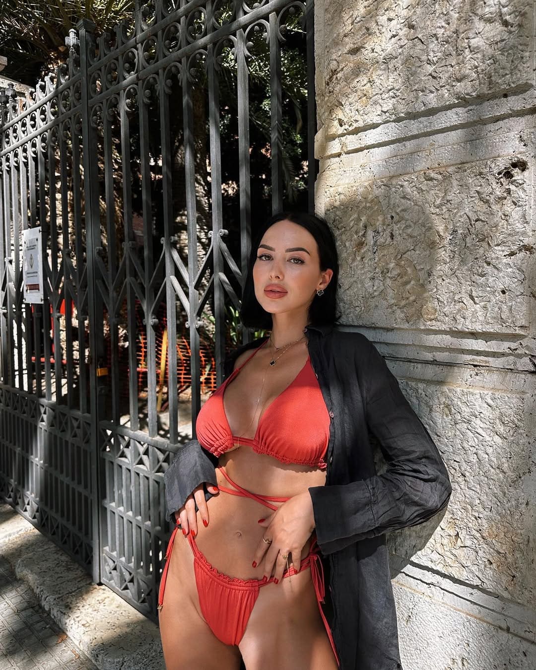 Mujer de bikini rojo y camisa oscura sobre hombros apoyada en muro de piedra rústica.