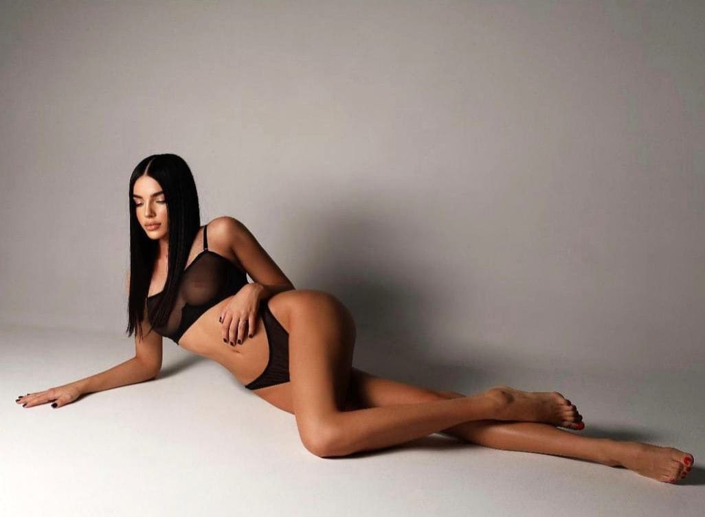 Donna sdraiata di lato su pavimento bianco in reggiseno e slip neri trasparenti, un braccio esteso in avanti.