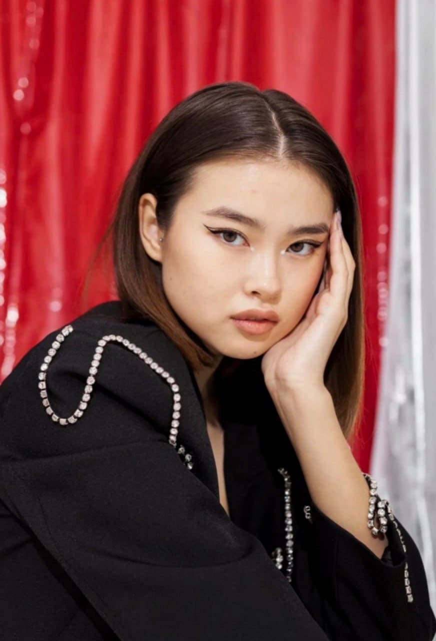 A woman with middle-parted hair rests her chin on her hand, wearing a crystal-trimmed jacket before red curtains.