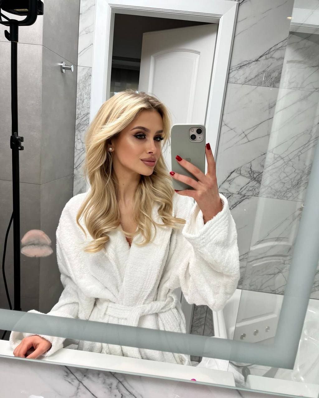 A blonde woman takes a mirror selfie in a marble bathroom wearing a white waffle-knit robe.