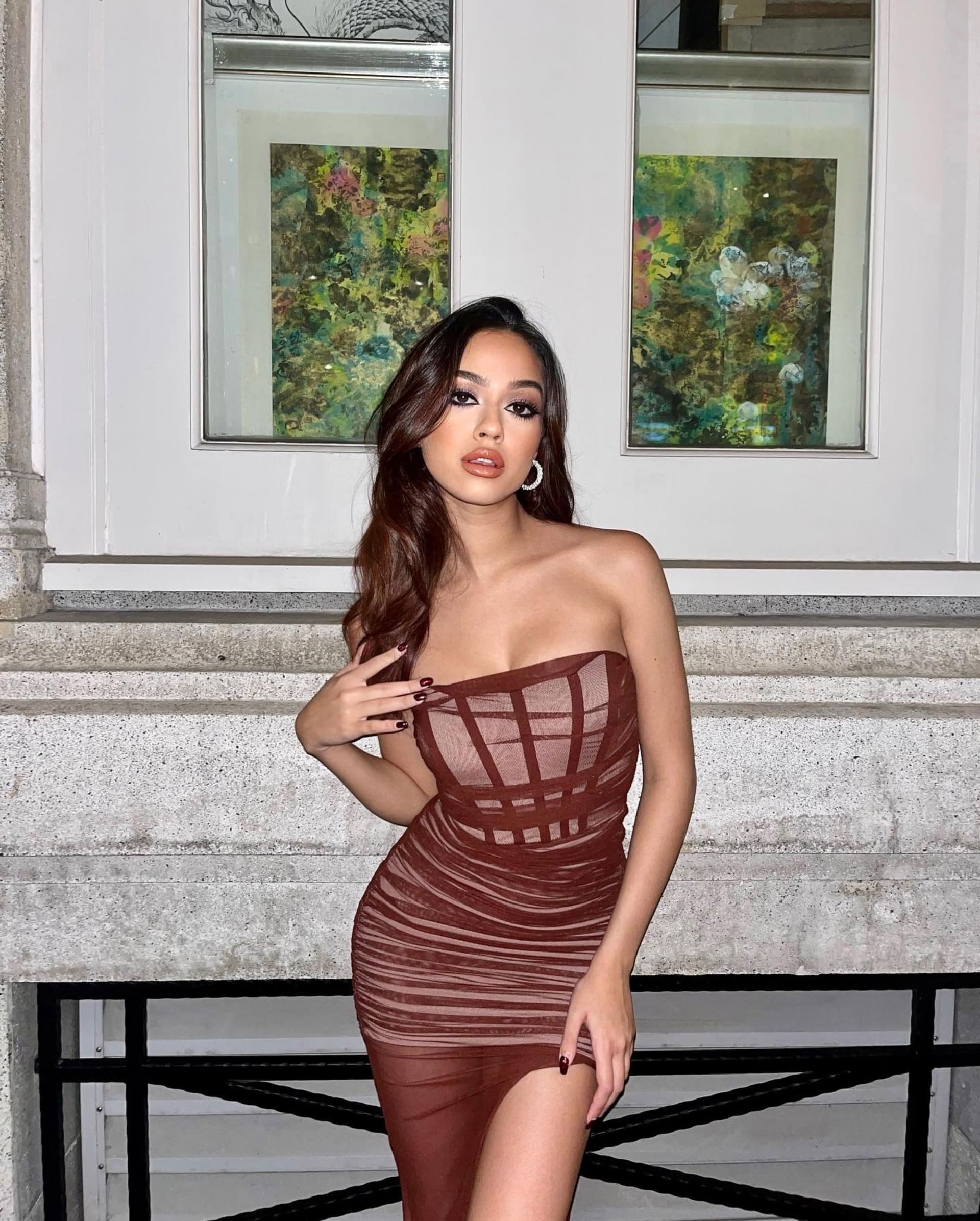 A woman in a brown slit dress poses against a wall with two framed paintings.