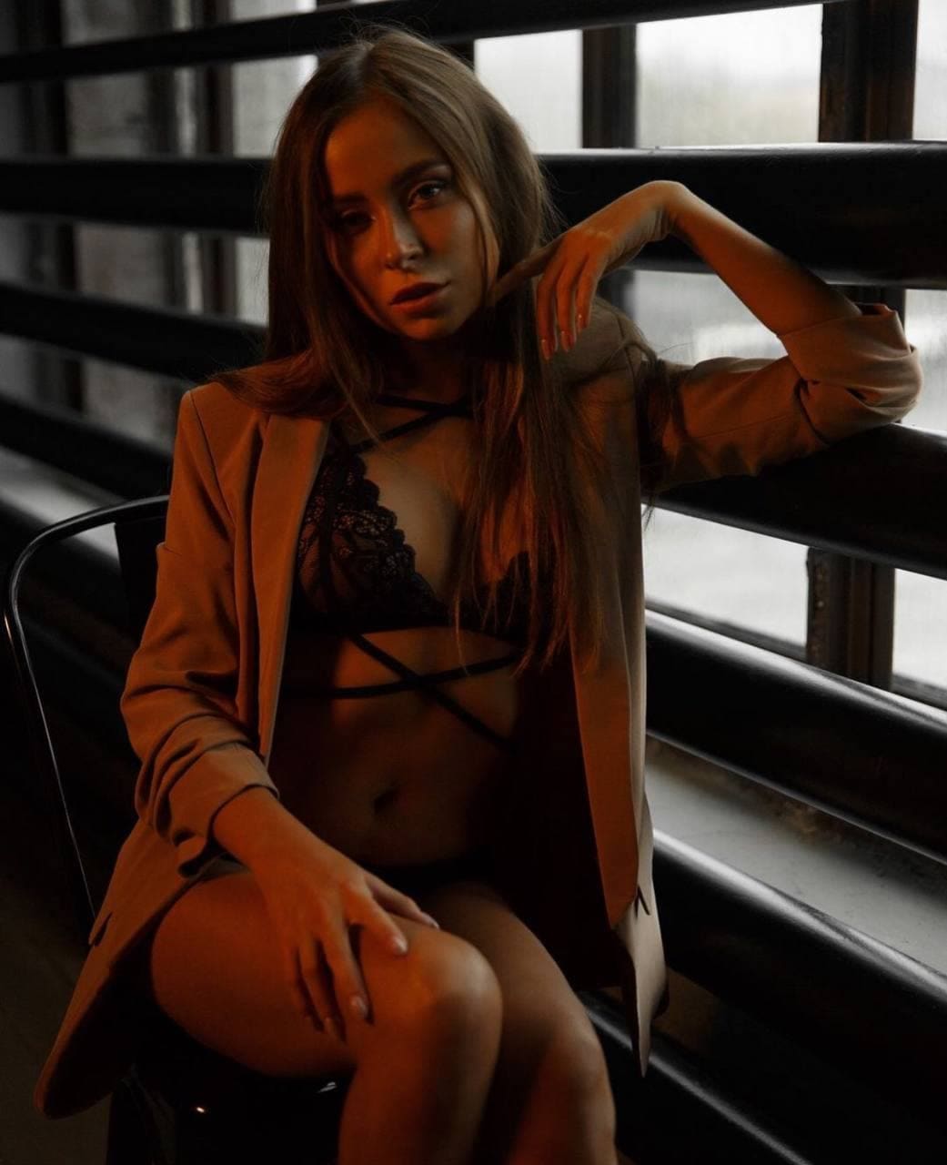 Donna seduta su una sedia davanti a una ringhiera metallica scura, con blazer color sabbia aperto sopra un reggiseno in pizzo nero.