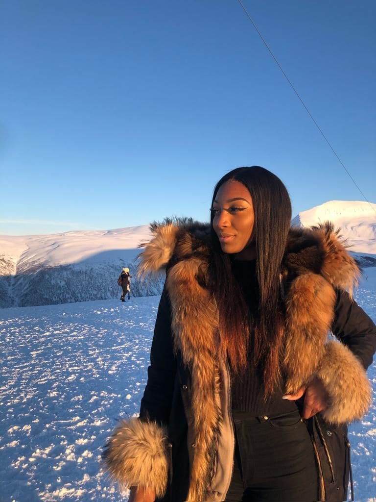 Donna in montagna innevata con cappotto nero dal grande colletto di pelliccia marrone, cielo azzurro alle spalle.