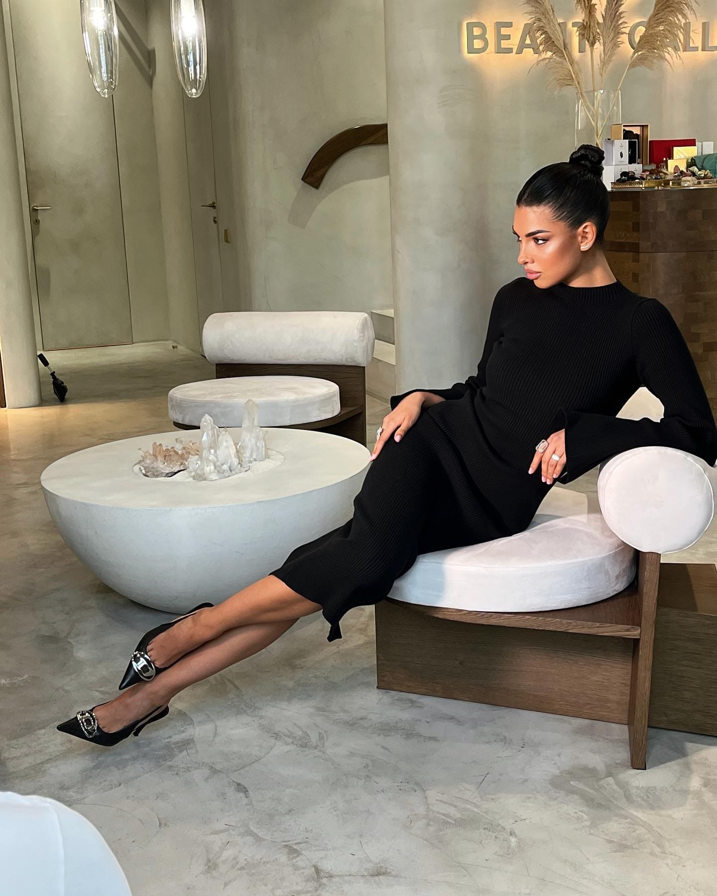 Mujer con moño alto sentada en un banco acolchado blanco en un interior moderno, vistiendo un vestido negro de punto y tacones negros de punta, con las piernas cruzadas.