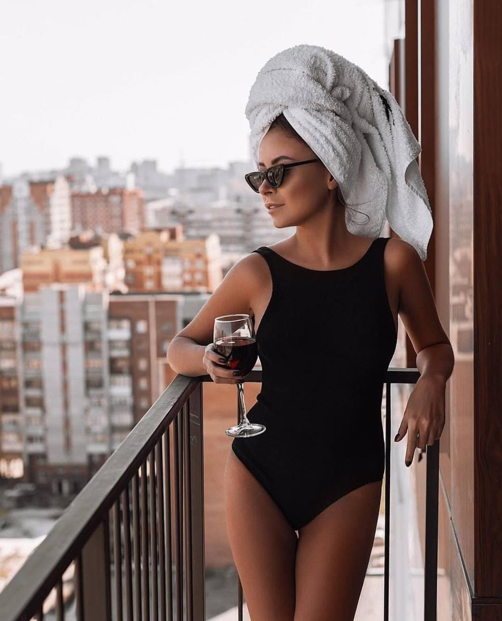 Donna con asciugamano bianco in testa e occhiali da sole in piedi su un balcone, tiene un bicchiere di vino rosso, sfondo urbano.