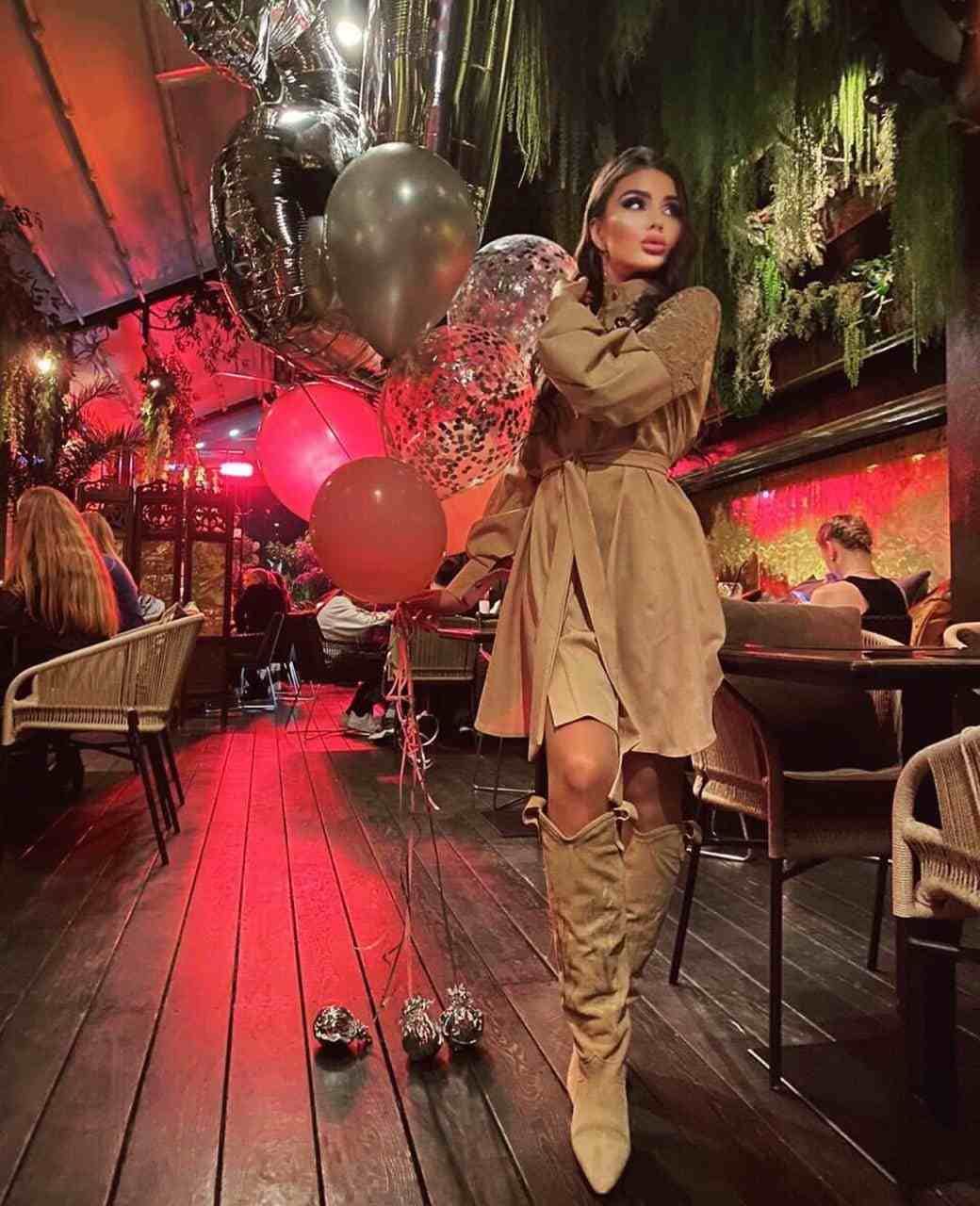 A woman stands on a wooden deck in a tan belted dress and suede boots, holding red and silver balloons in a dimly lit restaurant.
