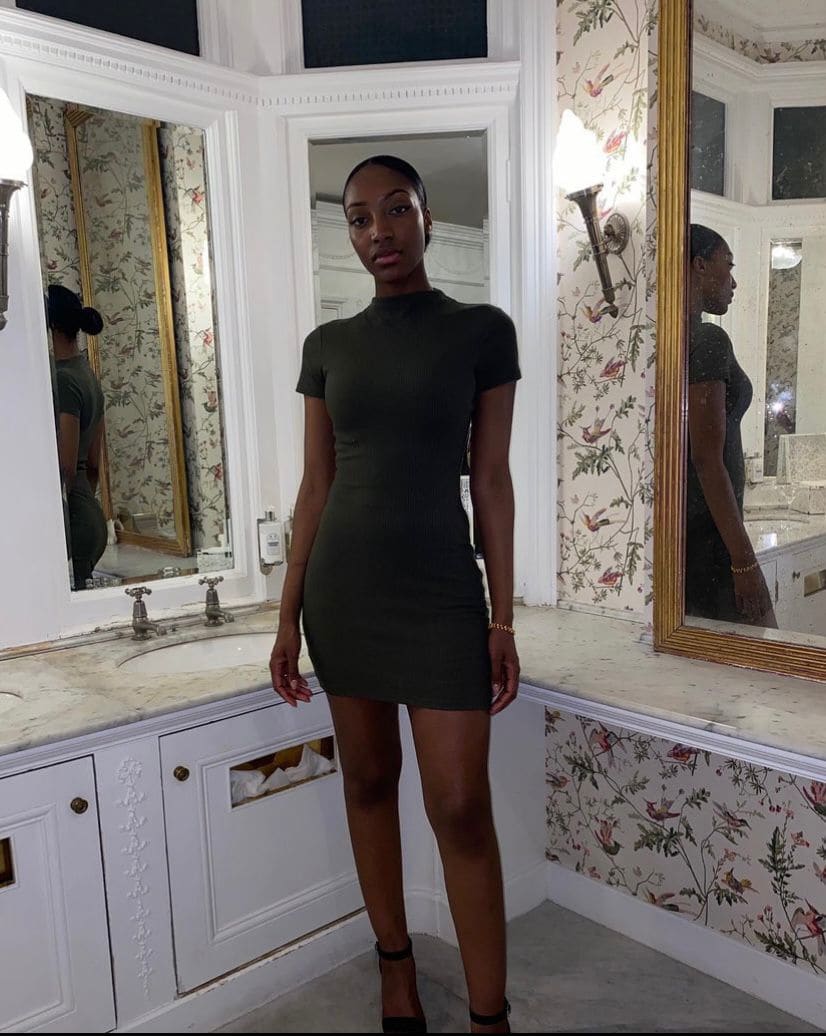 Donna con capelli scuri raccolti in un abito verde oliva corto, in bagno con carta da parati floreale e mobili bianchi.