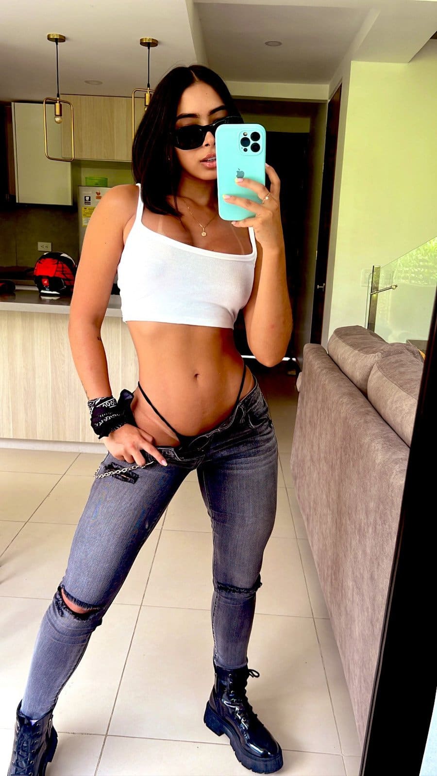 Mujer con gafas de sol y top blanco recortado tomando una selfie frente a un espejo en una cocina moderna.