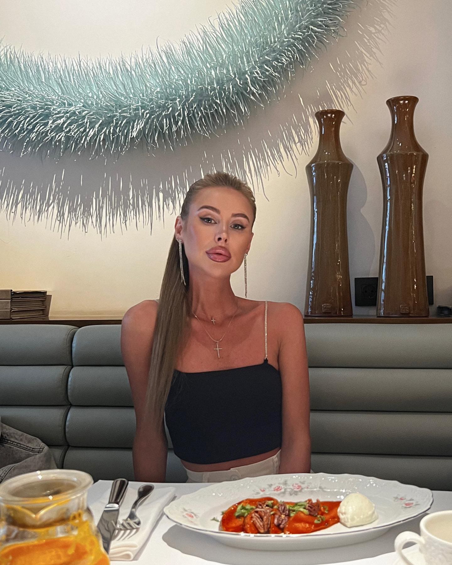 Tamilа sitzt am Restauranttisch in schwarzem Top und weißer Hose vor Pasta und Saft.