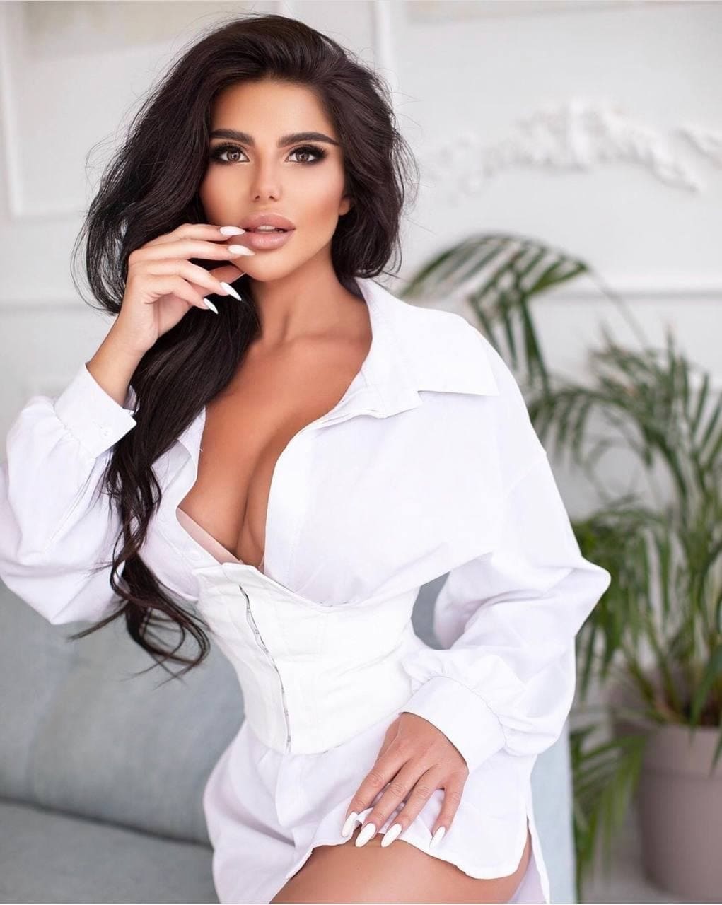 Anait in a white open shirt and corset poses with one hand near her chin in a bright room with a plant.