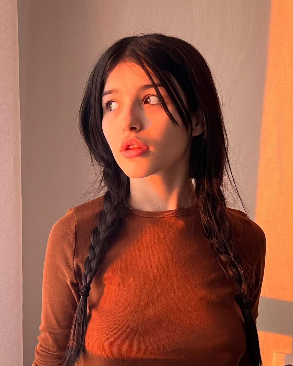Disha con dos trenzas sueltas y flequillo, viste una camiseta marrón de manga larga mirando hacia arriba en una habitación con luz cálida.