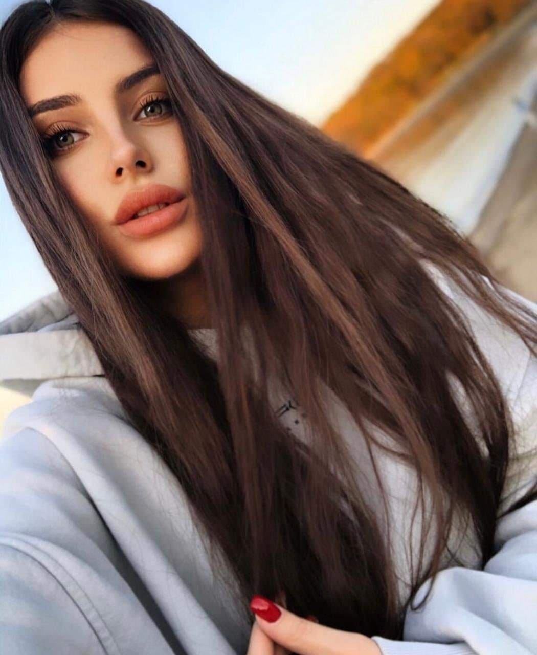 Close-up of a woman in a light grey hoodie with red nail polish, one hand near her chest, against a blurred warm outdoor backdrop.