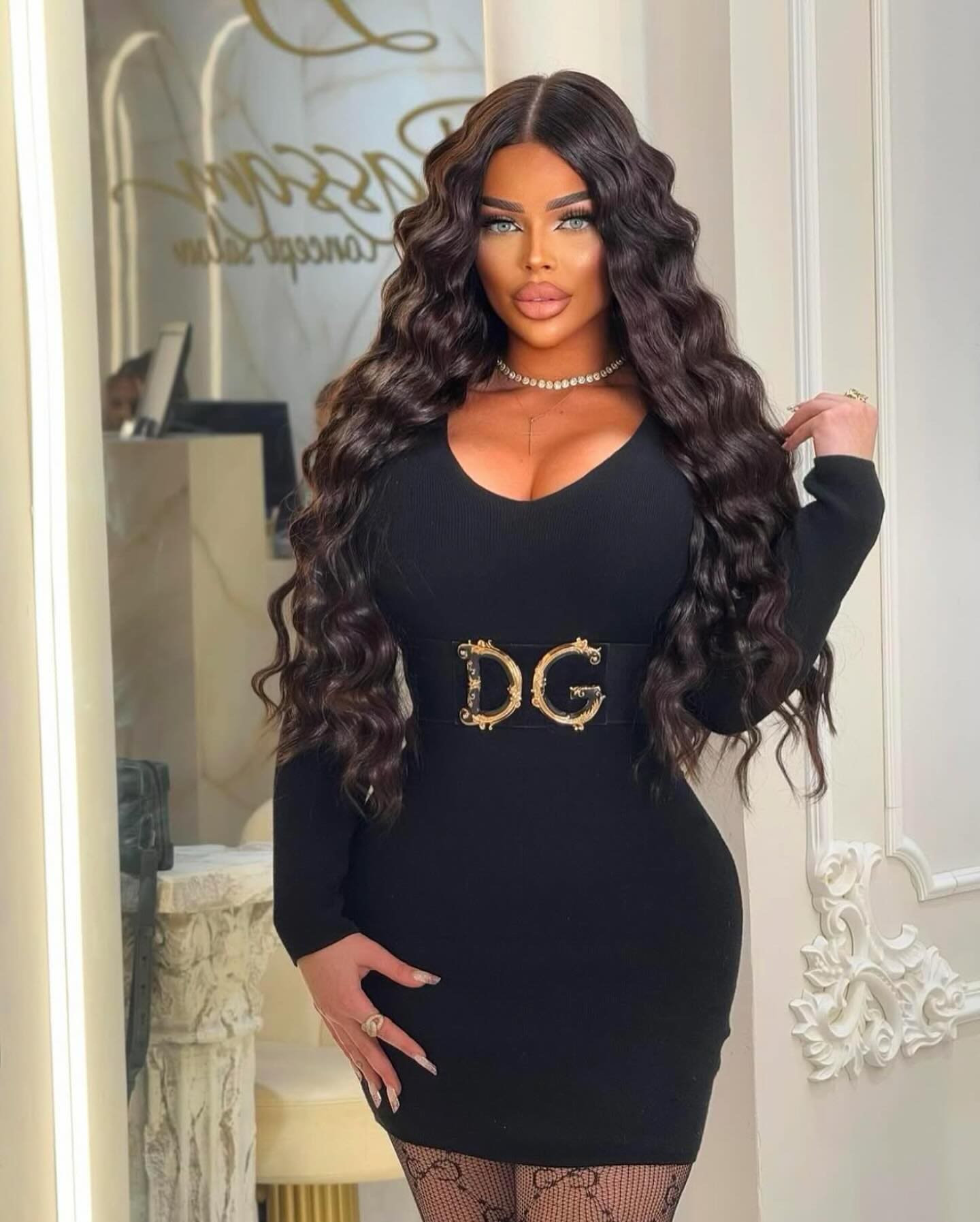 Mujer de cabello oscuro en vestido negro mini con escote V, cinturón dorado y medias de red.