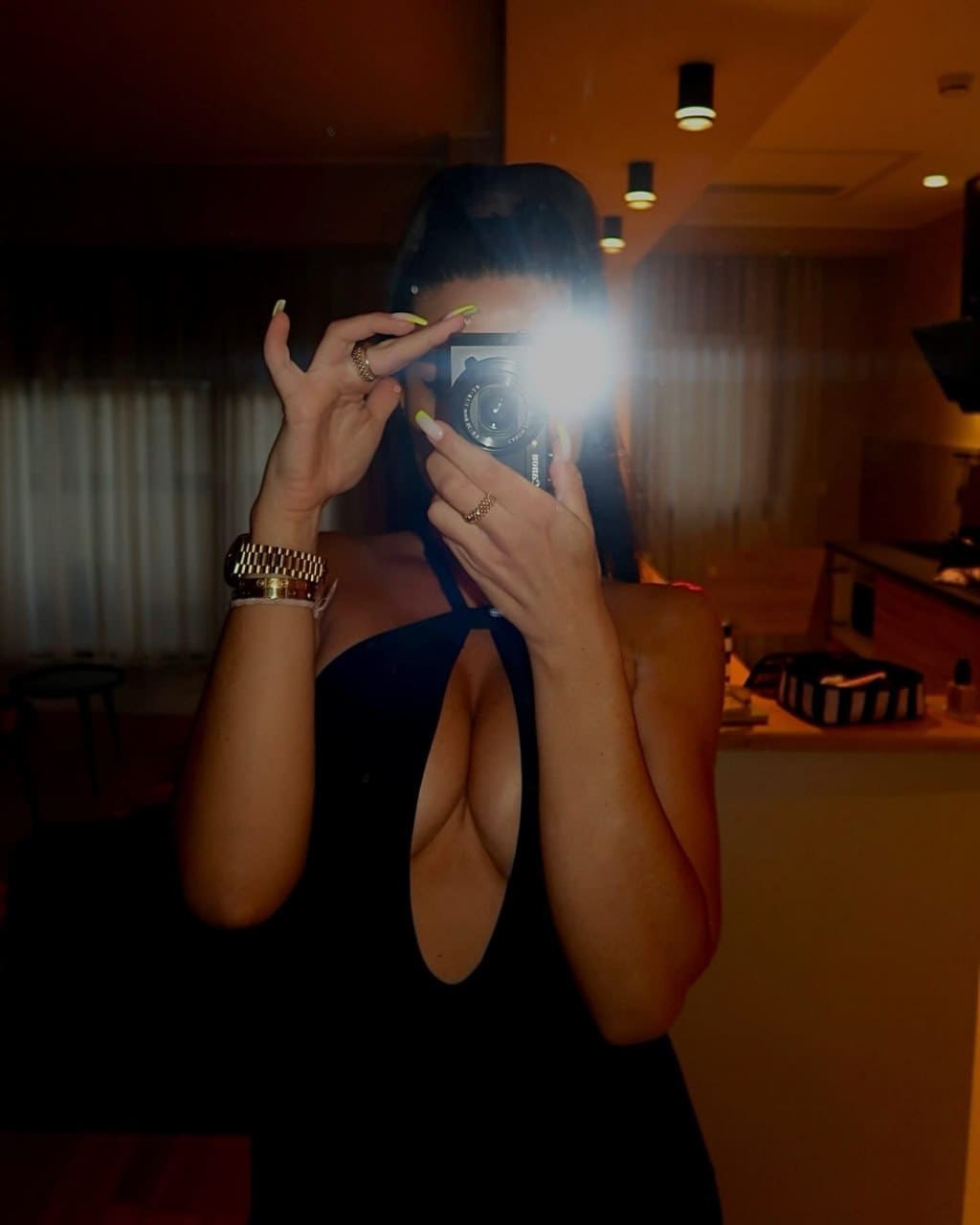 Autoportrait au miroir, visage masqué par le flash, haut noir sans manches et montre dorée.