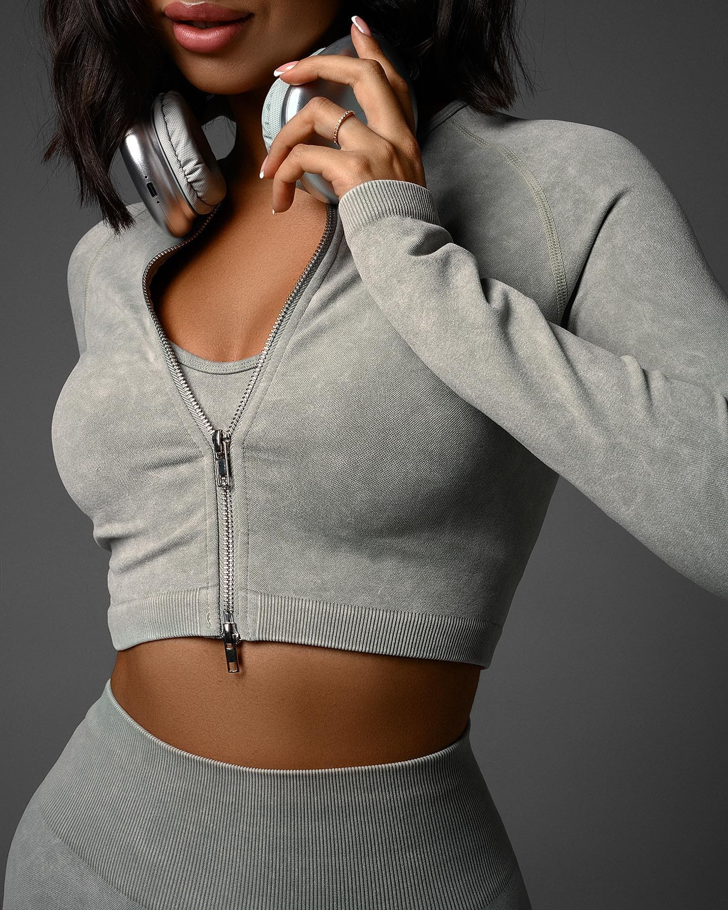 Mujer con cabello oscuro hasta los hombros usando sudadera corta gris con cremallera y leggings a juego, con auriculares plateados alrededor del cuello y una mano cerca de la barbilla.