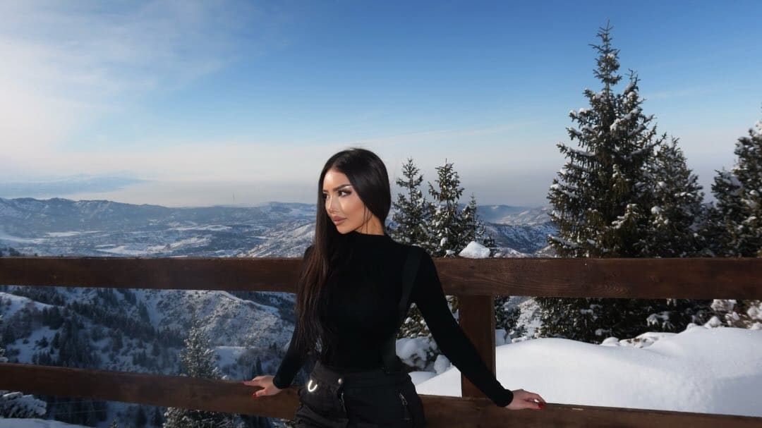 Mujer de pie en una barandilla de madera en una montaña nevada, con cuello alto negro de manga larga y pantalones oscuros, mirando hacia el paisaje.