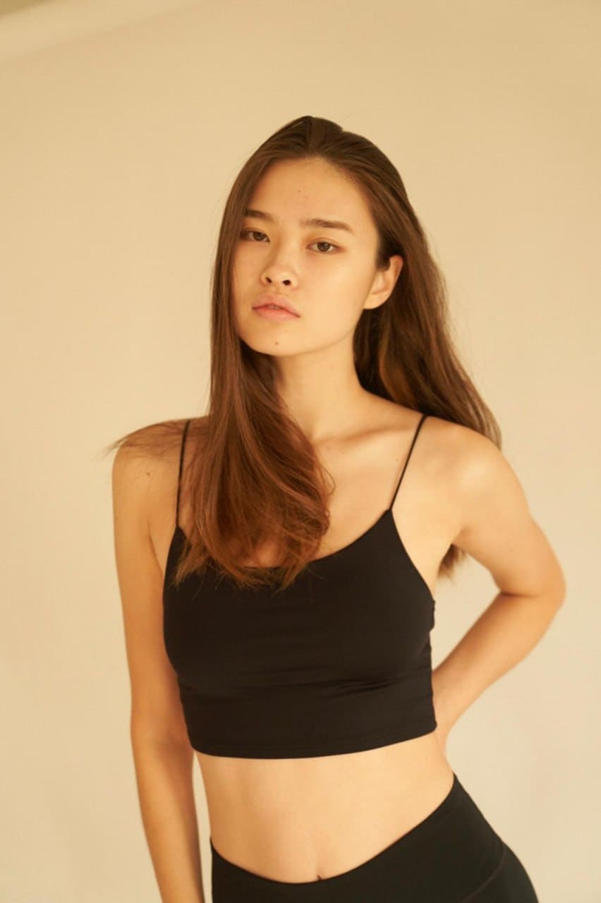 A young woman with long brown hair stands against a beige wall in a black crop top and high-waisted bottoms.