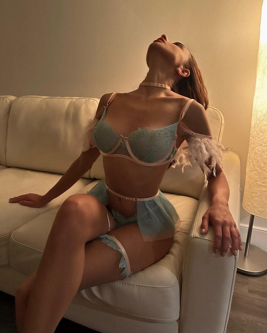Mujer de pelo castaño en sofá de cuero crema, con lencería azul clara y detalles de plumas, cabeza inclinada hacia atrás.