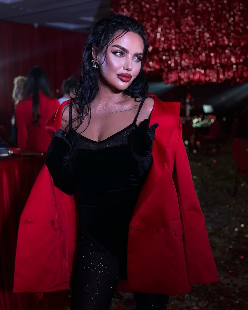 Mujer de pie en sala oscura, vestido negro de terciopelo y blazer rojo sobre hombros.