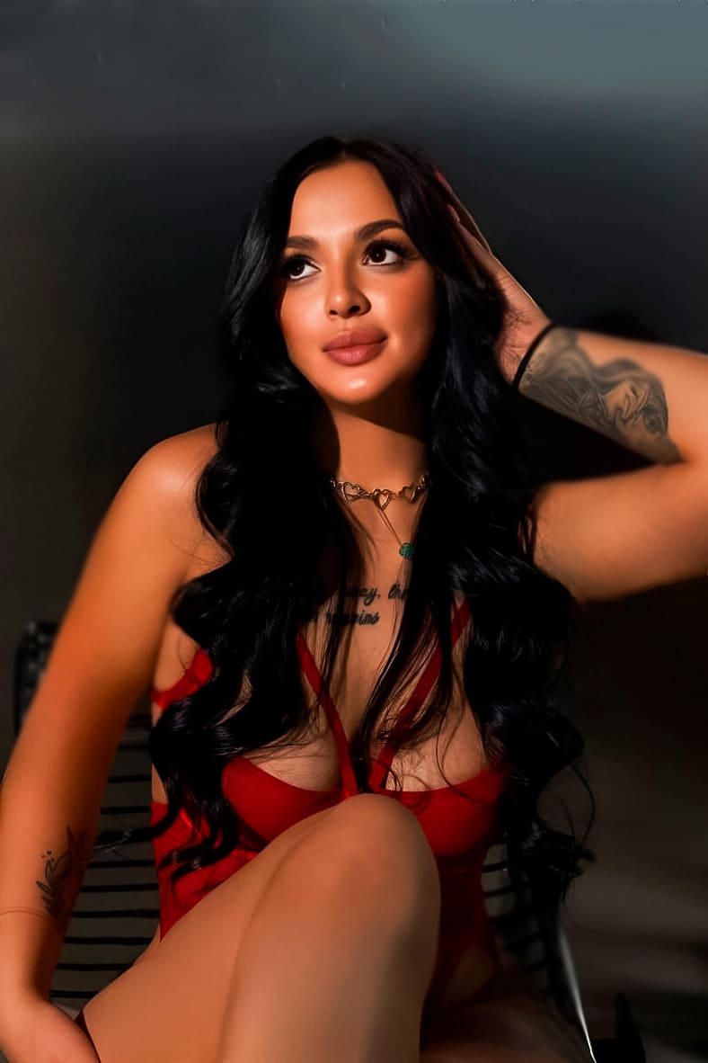 Mujer sentada en silla con bodysuit rojo de tirantes, una mano levantada hacia la cabeza contra un fondo oscuro.