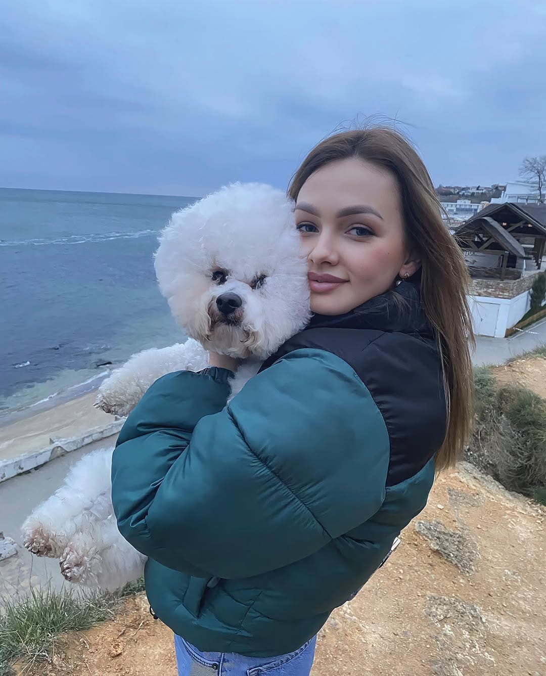 Mujer morena sosteniendo un perro blanco peludo en una ladera hierbosa frente al agua, con chaqueta verde y negra y vaqueros azules.