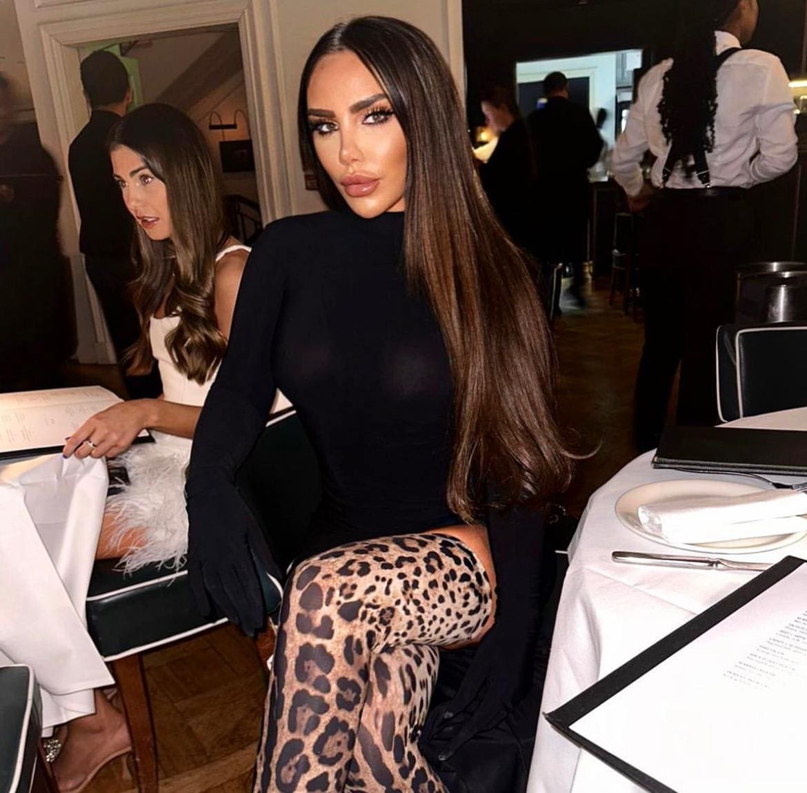 A woman sits at a restaurant table in a black long-sleeved top and leopard-print tights, with a menu and white cloth in front.