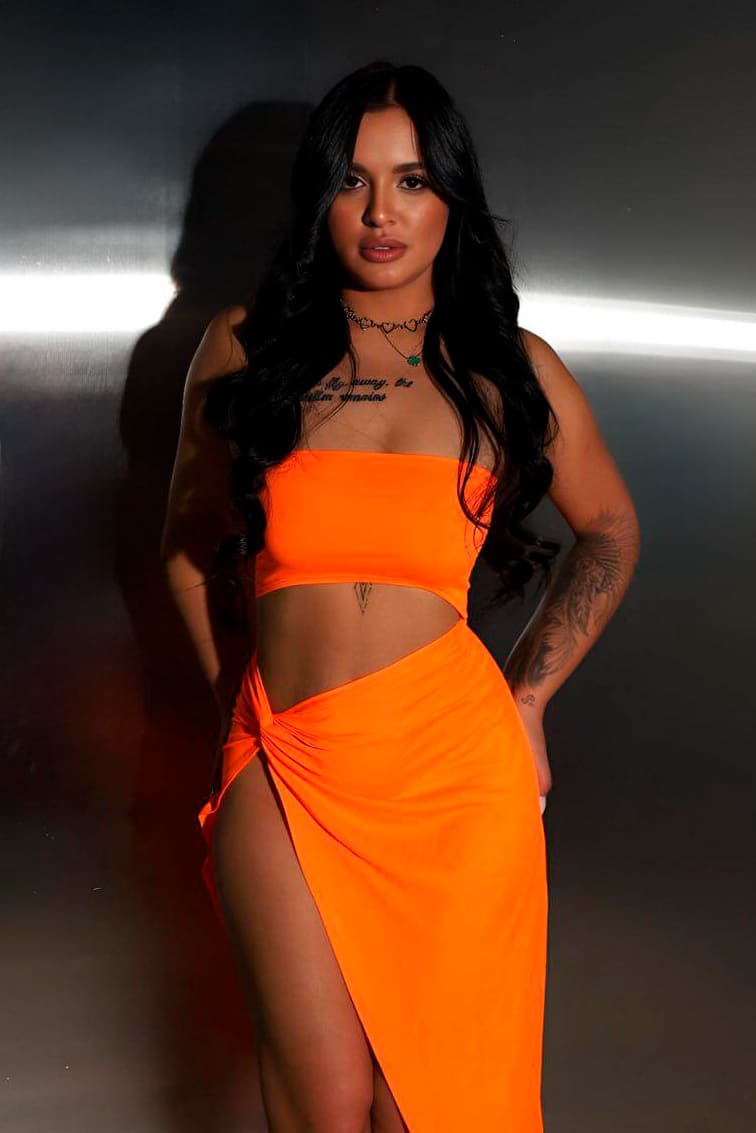 Mujer de pie con conjunto naranja sin tirantes y falda aberturas, tatuaje visible en brazo y gargantilla al cuello.