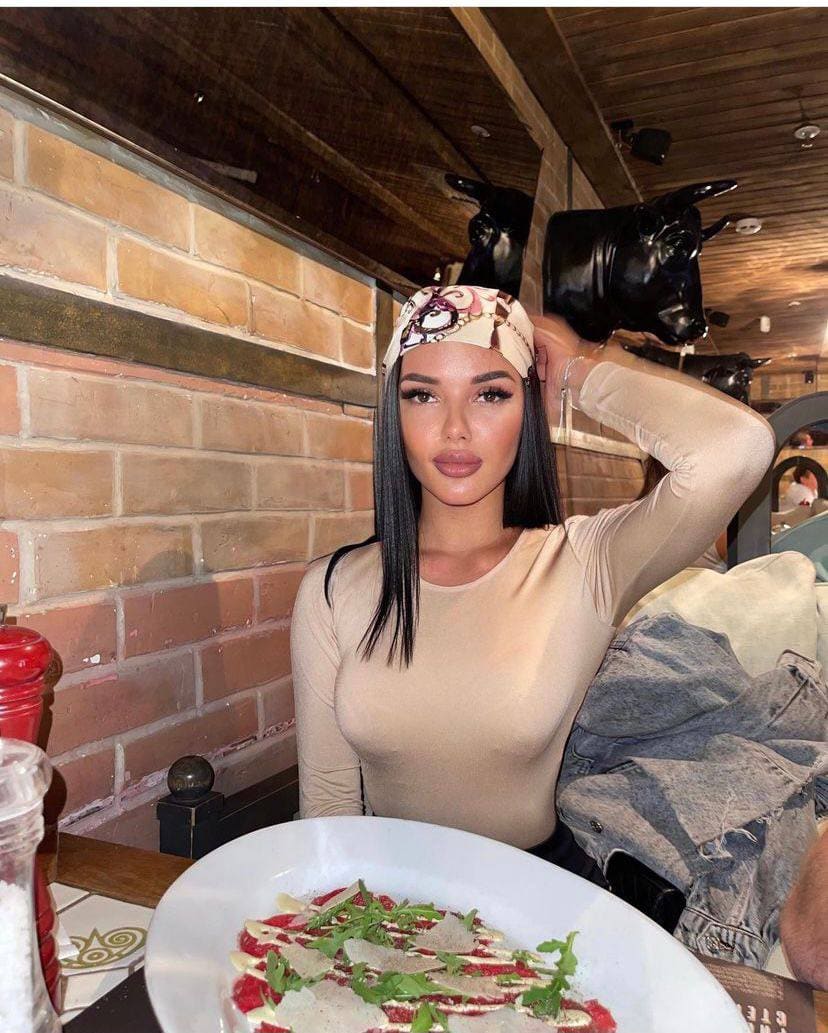 Mujer con pañuelo estampado y top beige de manga larga sentada en mesa de restaurante con plato de comida y mano junto a la cabeza.