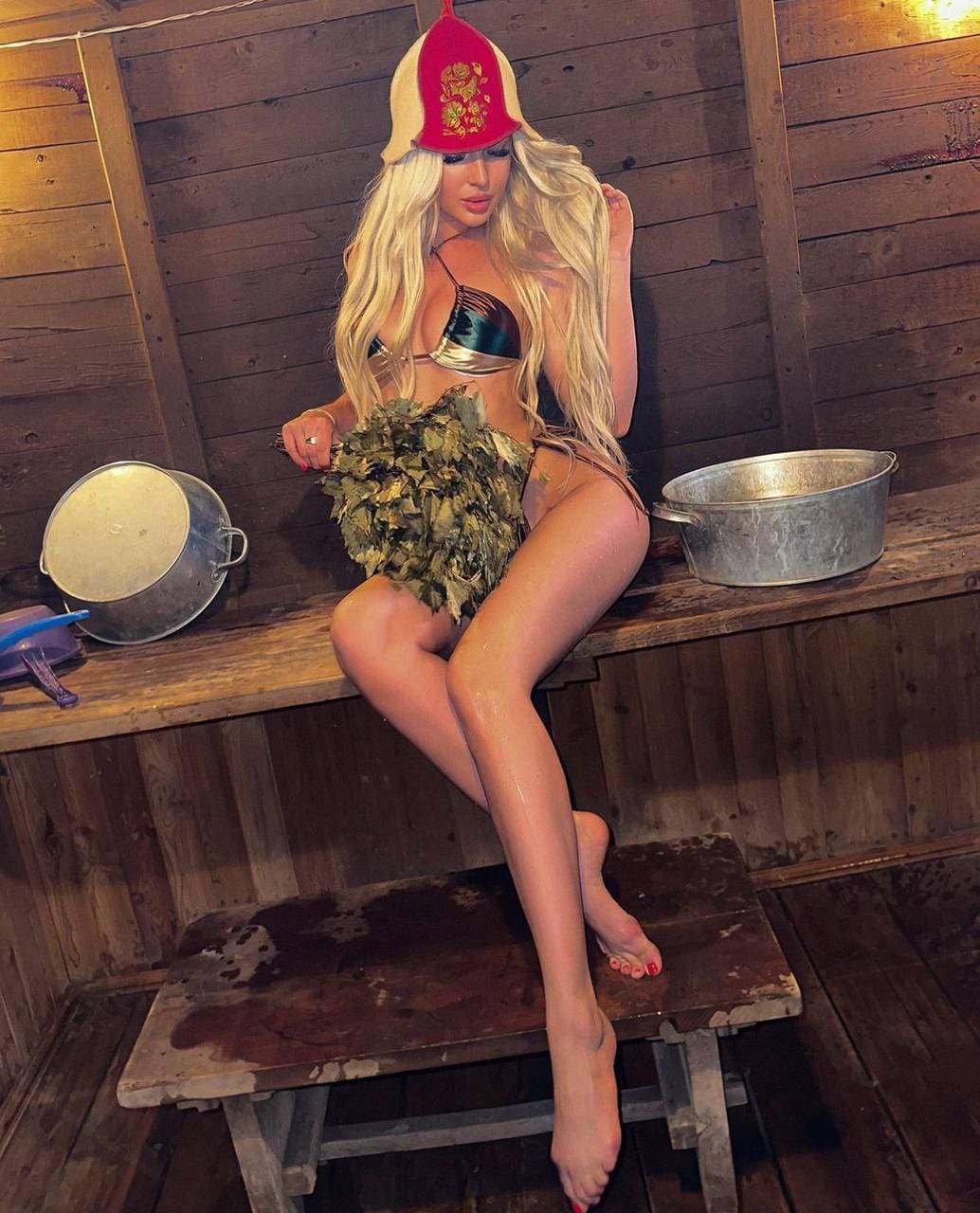 A woman sits on a wooden bench in a rustic room, wearing an embroidered hat, metallic bikini top, and leafy skirt.