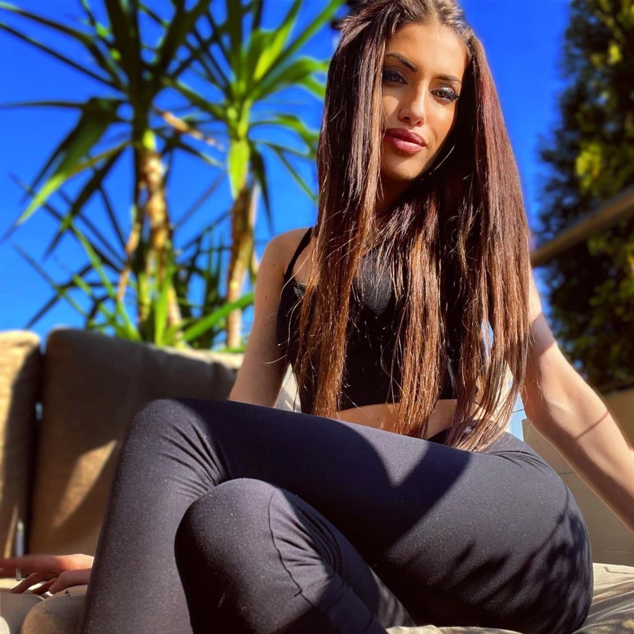 Donna con top e leggings neri, seduta su panca esterna all'aperto tra palme e cielo azzurro.