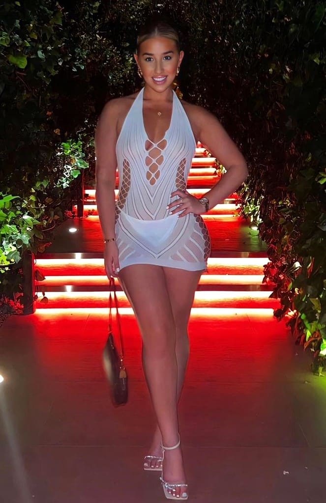 Anissa con vestido blanco de tirantes y aberturas, sosteniendo un bolso en una escalera iluminada en rojo.