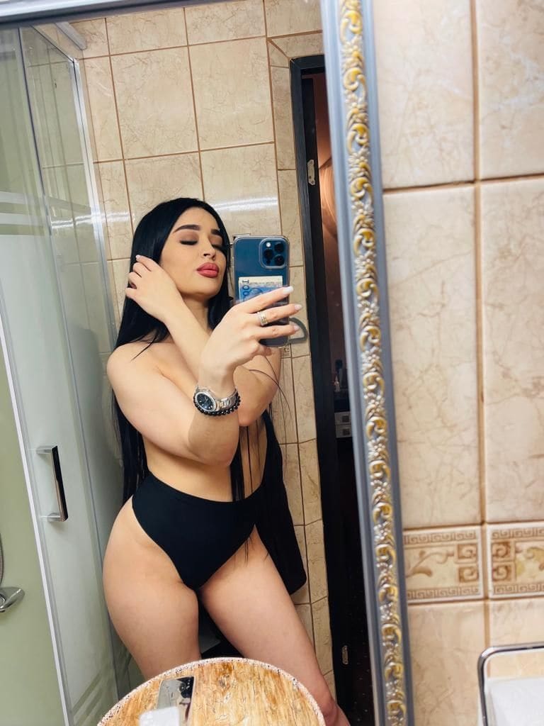 Donna con capelli scuri che si fa un selfie allo specchio in bagno piastrellato, indossa un costume intero nero e tiene un telefono blu.