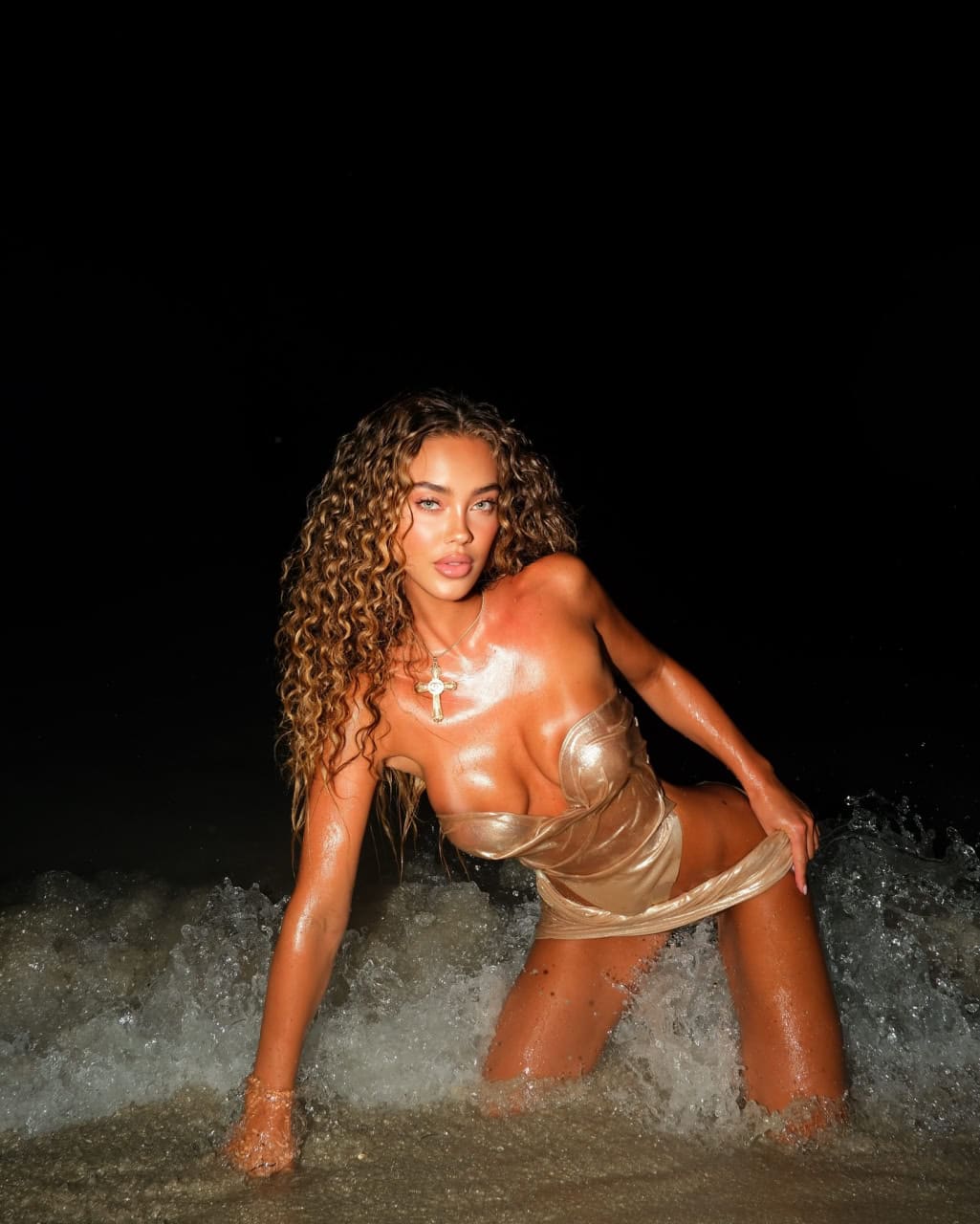 Mujer de cabello rizado arrodillada en el agua del océano nocturno, con vestido dorado sin tirantes y collar de cruz al cuello.