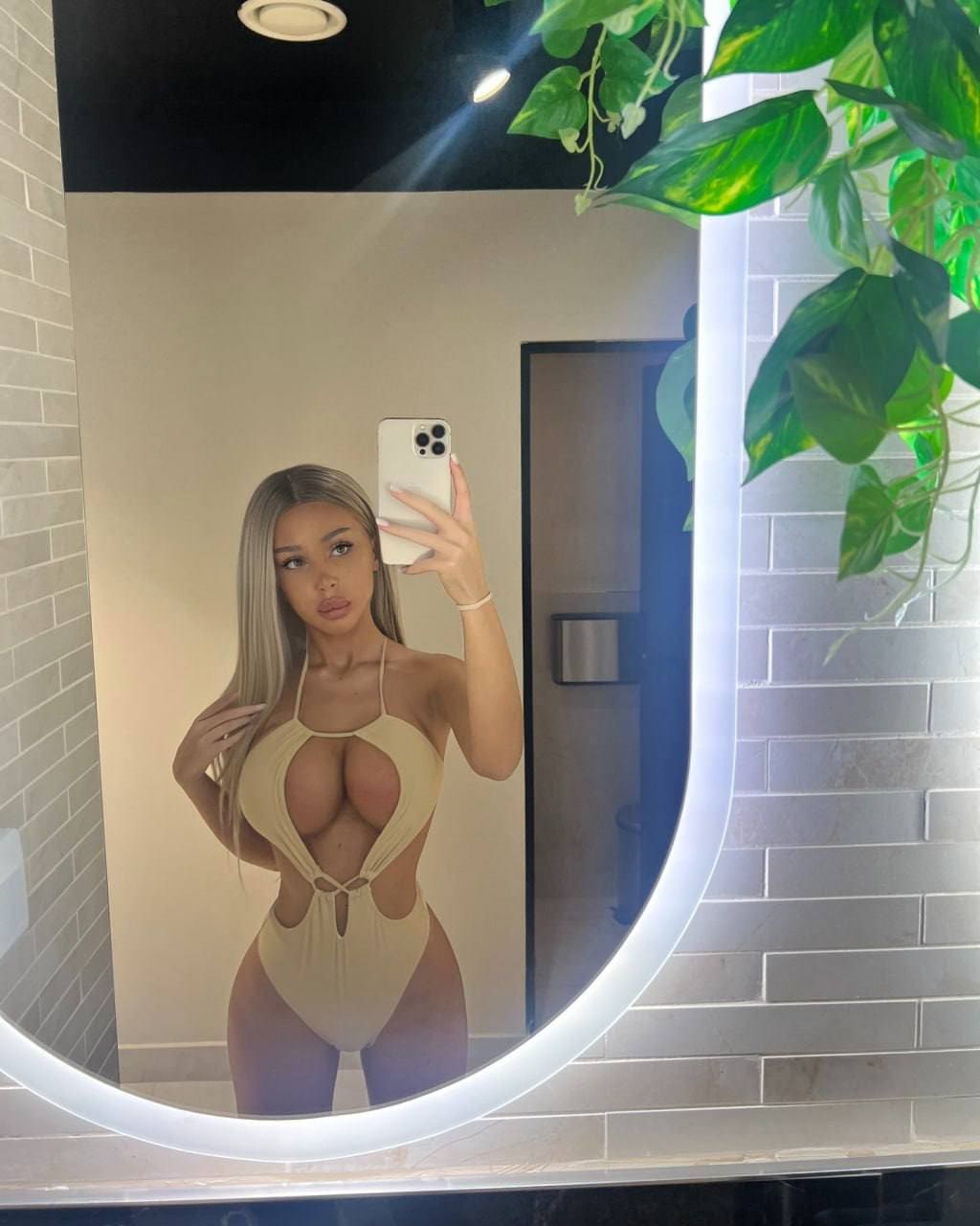 Mujer rubia tomando selfie en espejo con traje de baño beige de cortes, sosteniendo teléfono blanco, junto a planta verde.