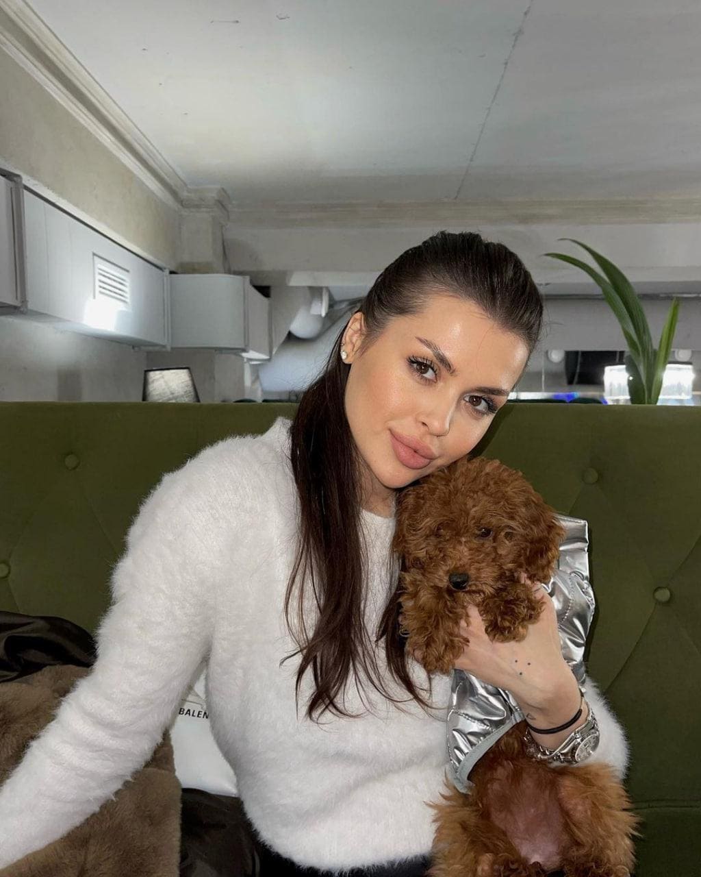 Mujer con cabello oscuro recogido sentada en un sofá verde tapizado, usando un suéter blanco peludo y sosteniendo un pequeño poodle marrón en el regazo mientras mira a la cámara.