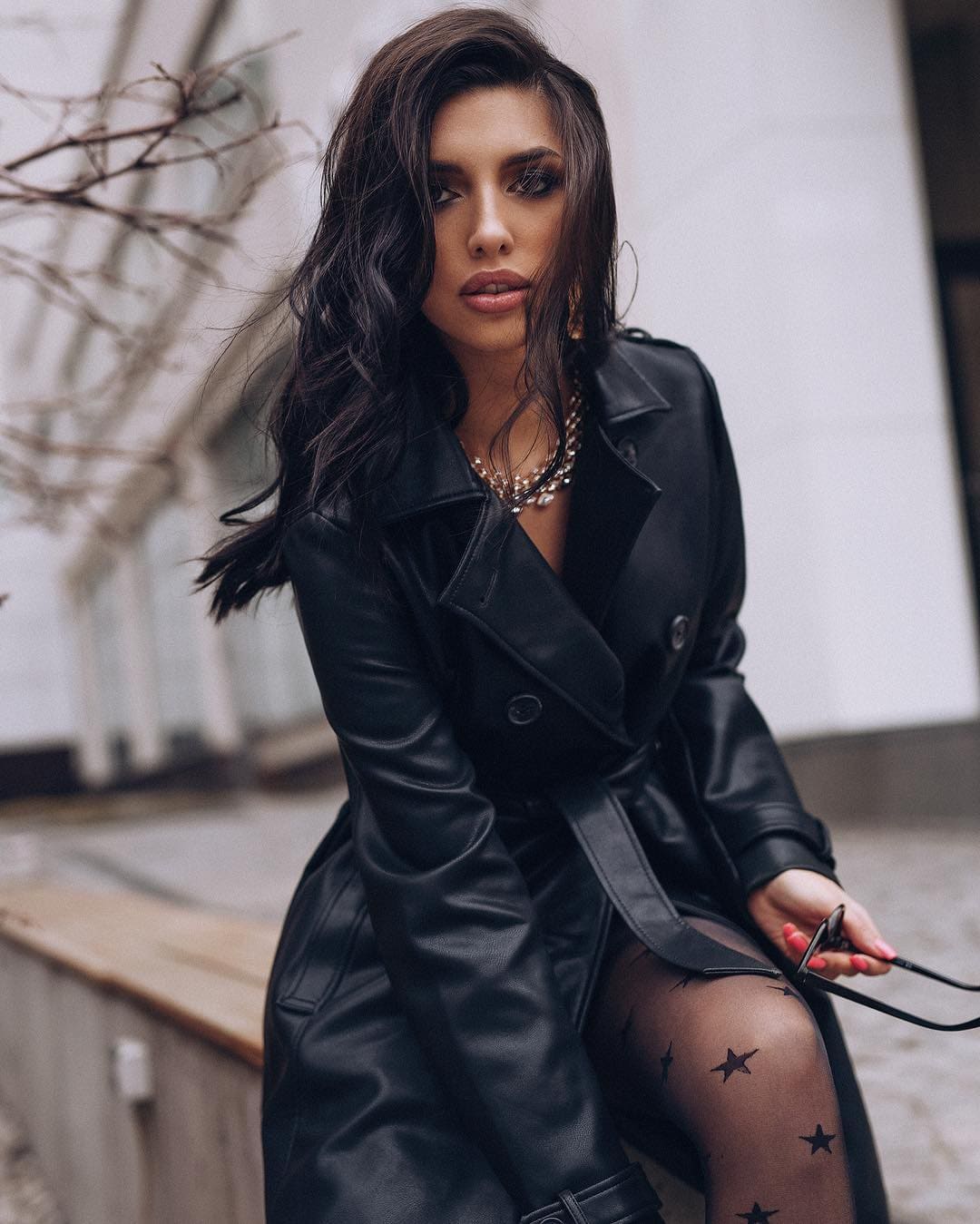 Mujer con cabello largo oscuro sentada en un banco de madera al aire libre, llevando un trench de cuero negro sobre medias negras con estrellas y sosteniendo unas gafas de sol.