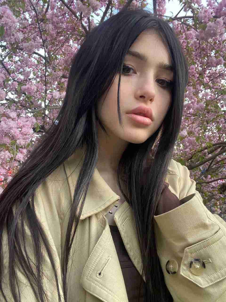 Mujer de cabello oscuro largo frente a árboles florales rosas, lleva un abrigo beige claro sobre una prenda oscura mirando a la cámara.